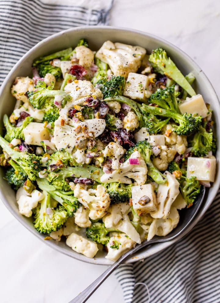 a large bowl filled with broccoli, cauliflower, cheese, and dried cranberries on top of a striped linen