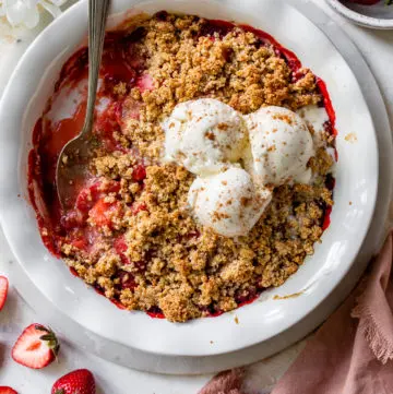strawberry crisp in a white pie pan topped with three scoops of vanilla ice cream