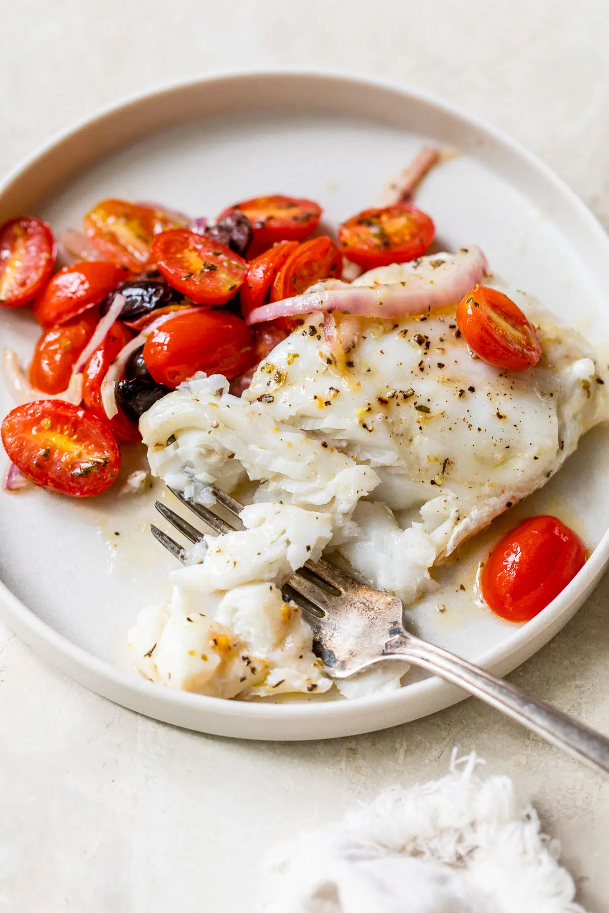 a piece of white fish on a plate beside cooked grape tomatoes
