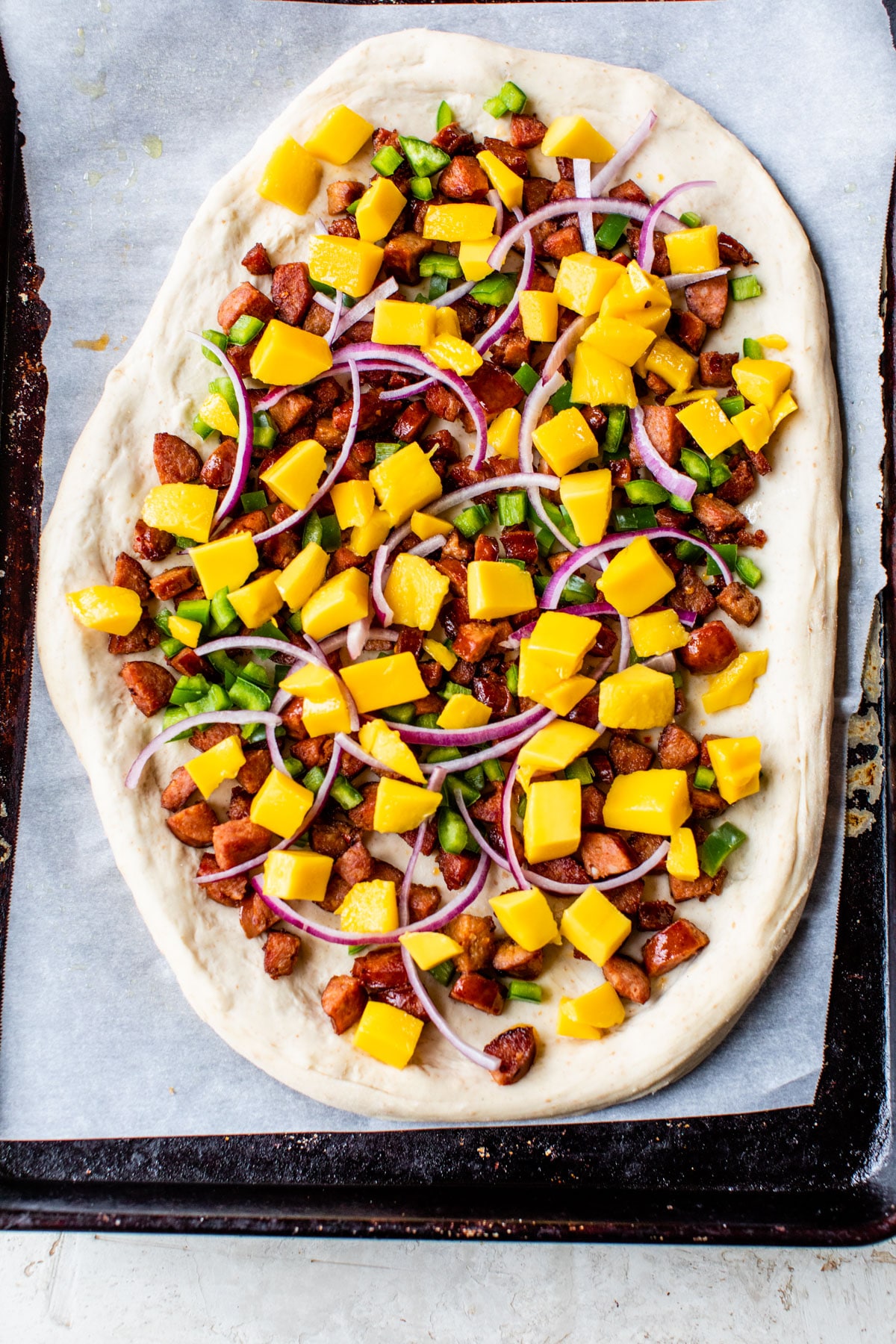uncooked pizza on a baking sheet topped with meat, mango, green pepper, and red onion