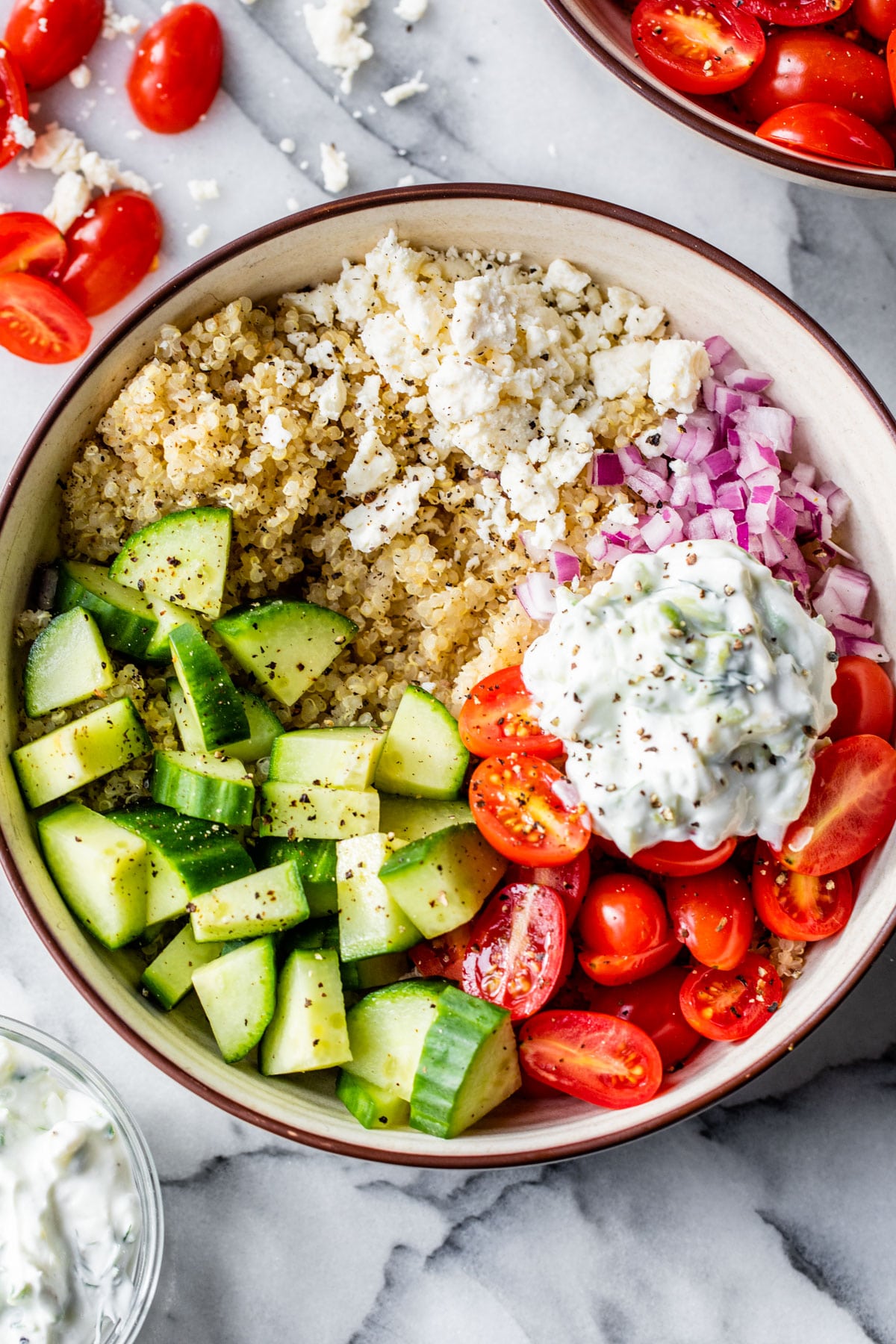 a bowl of quinoa topped with mediterranean ingredients like tomatoes, cucumber, red onion, and feta