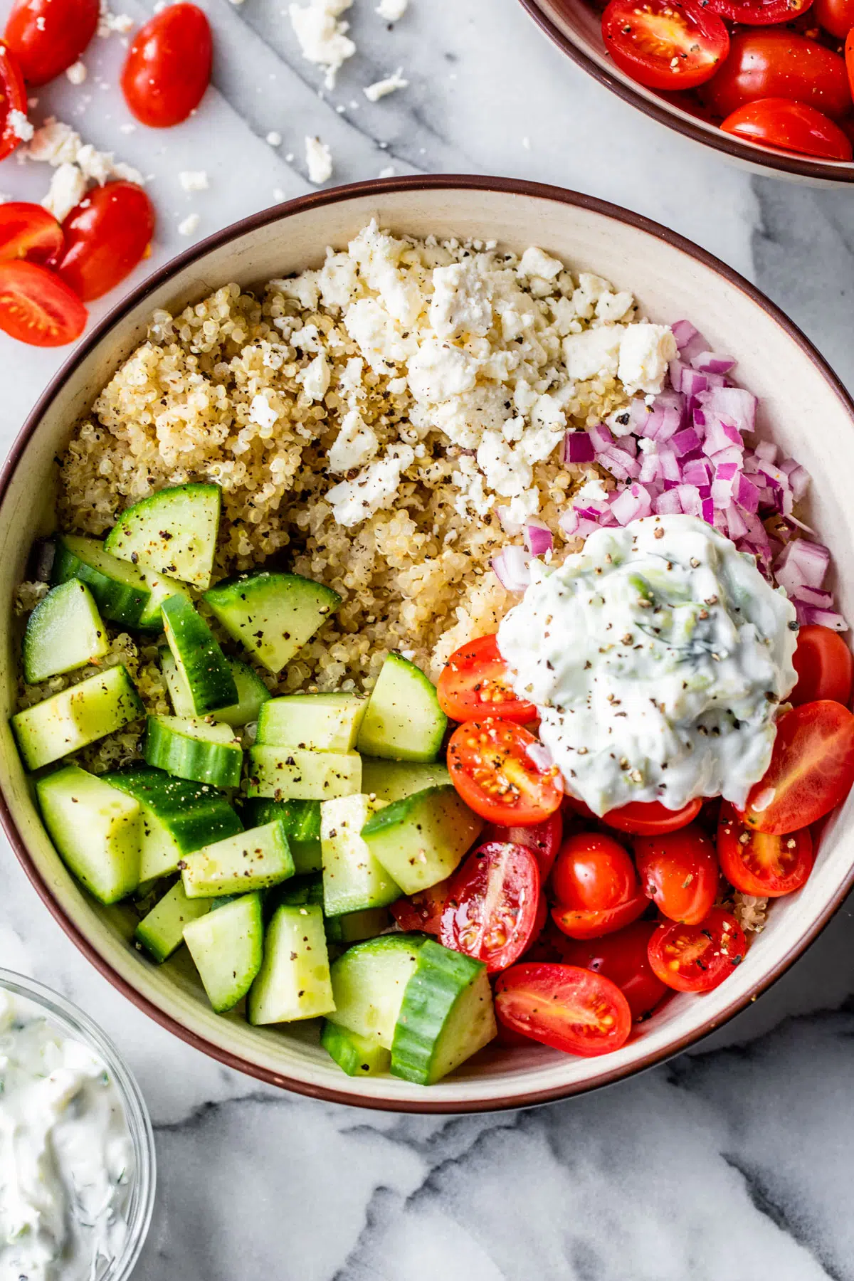 a bowl of quinoa topped with mediterranean ingredients like tomatoes, cucumber, red onion, and feta