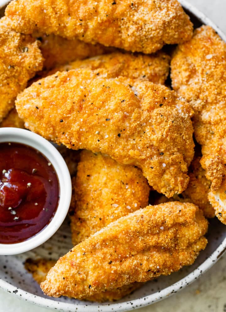 close up of breaded chicken tenders in a bowl beside a bowl of ketchup