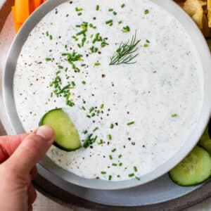 someone's hand dipping a cucumber into a bowl of white-colored dip