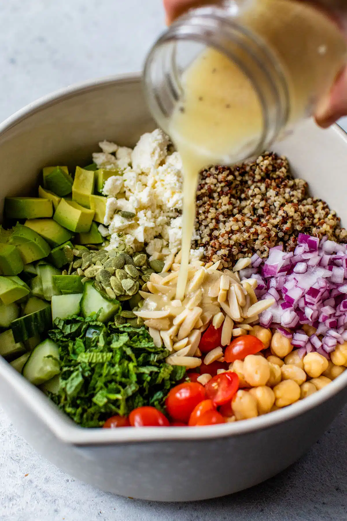 bowl with quinoa, feta cheese, cucumber, and other vegetables inside and dressing being poured overtop