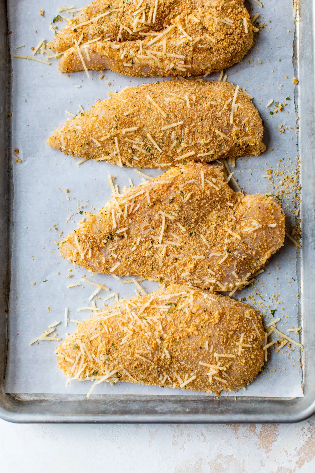 raw chicken breasts coated in cheese and breadcrumbs on a baking sheet