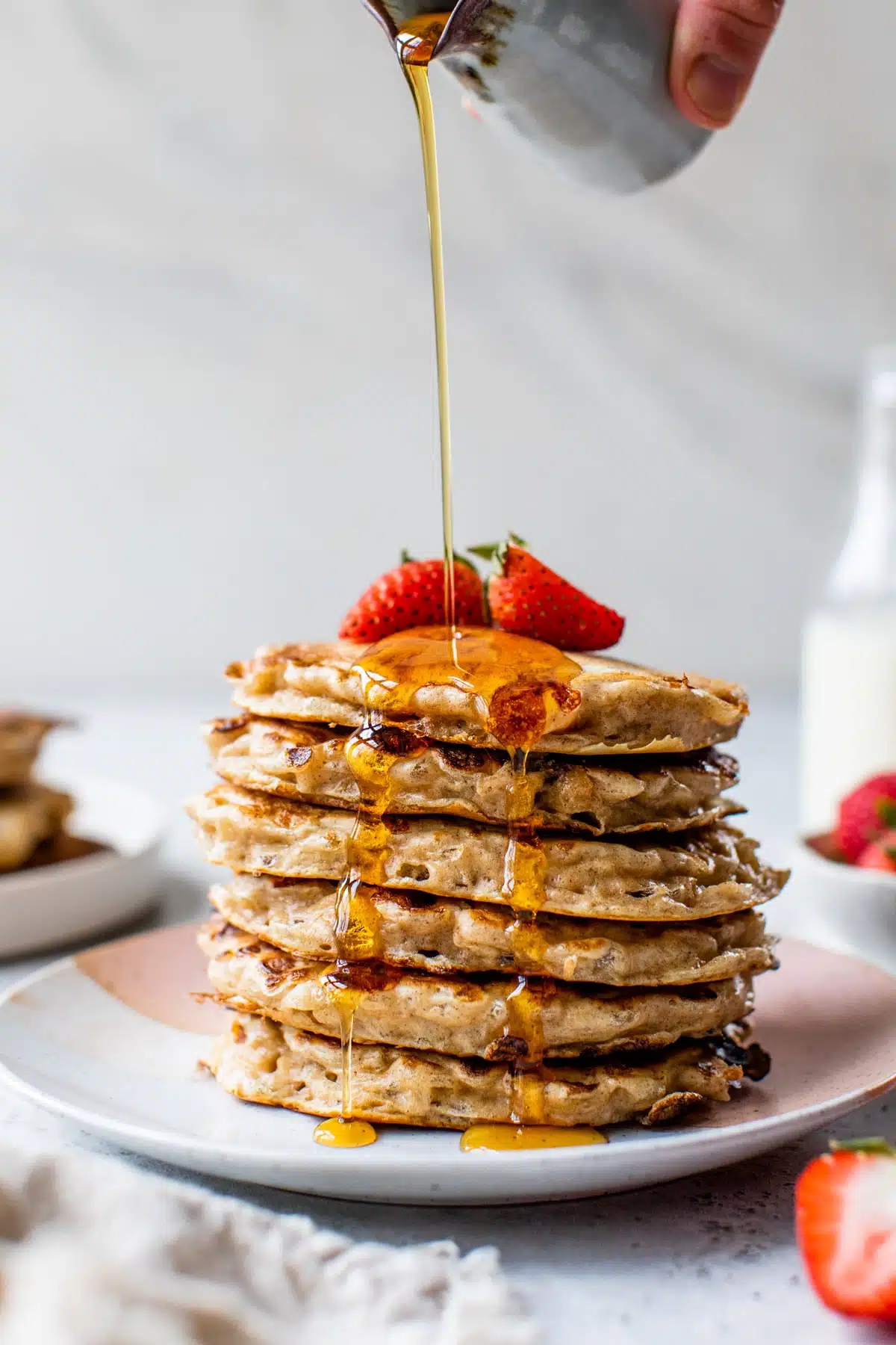 an side view of a stack of pancakes topped with two strawberries and maple syrup poured over the top