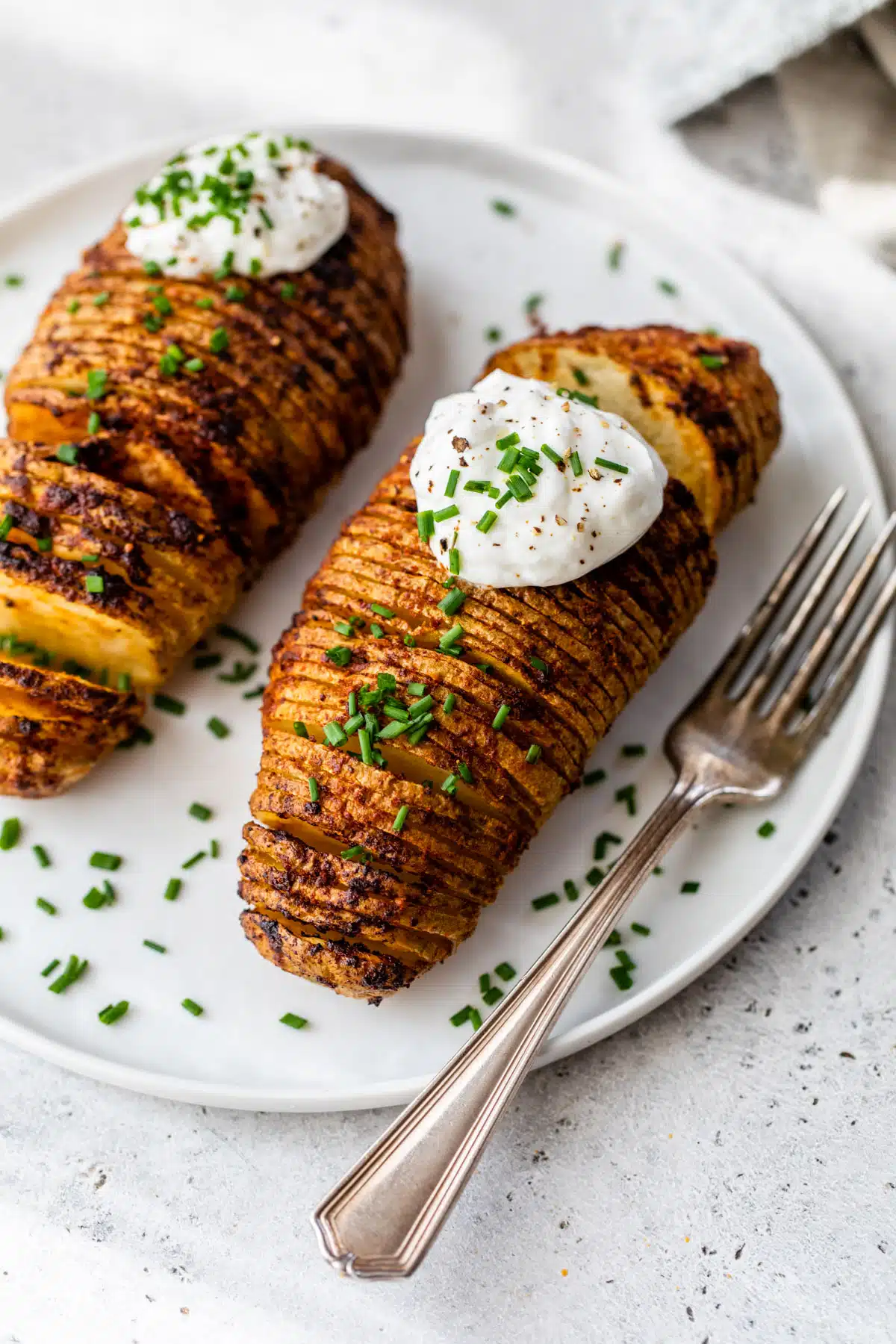 2 hasselback potatoes with sour cream and chives on top on a plate with a fork