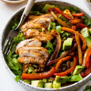 salad in a bowl topped with sliced chicken, bell peppers and diced avocado