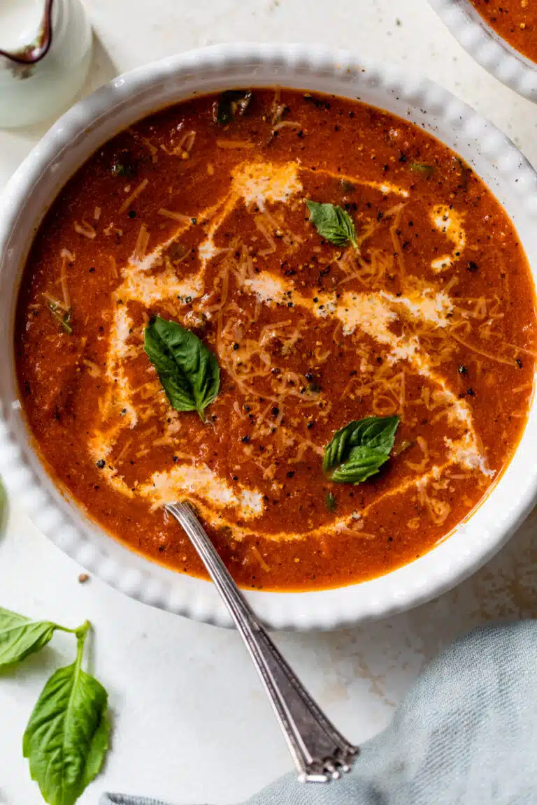 creamy tomato basil soup in a bowl with a spoon and with half & half drizzled in and basil leaves on top