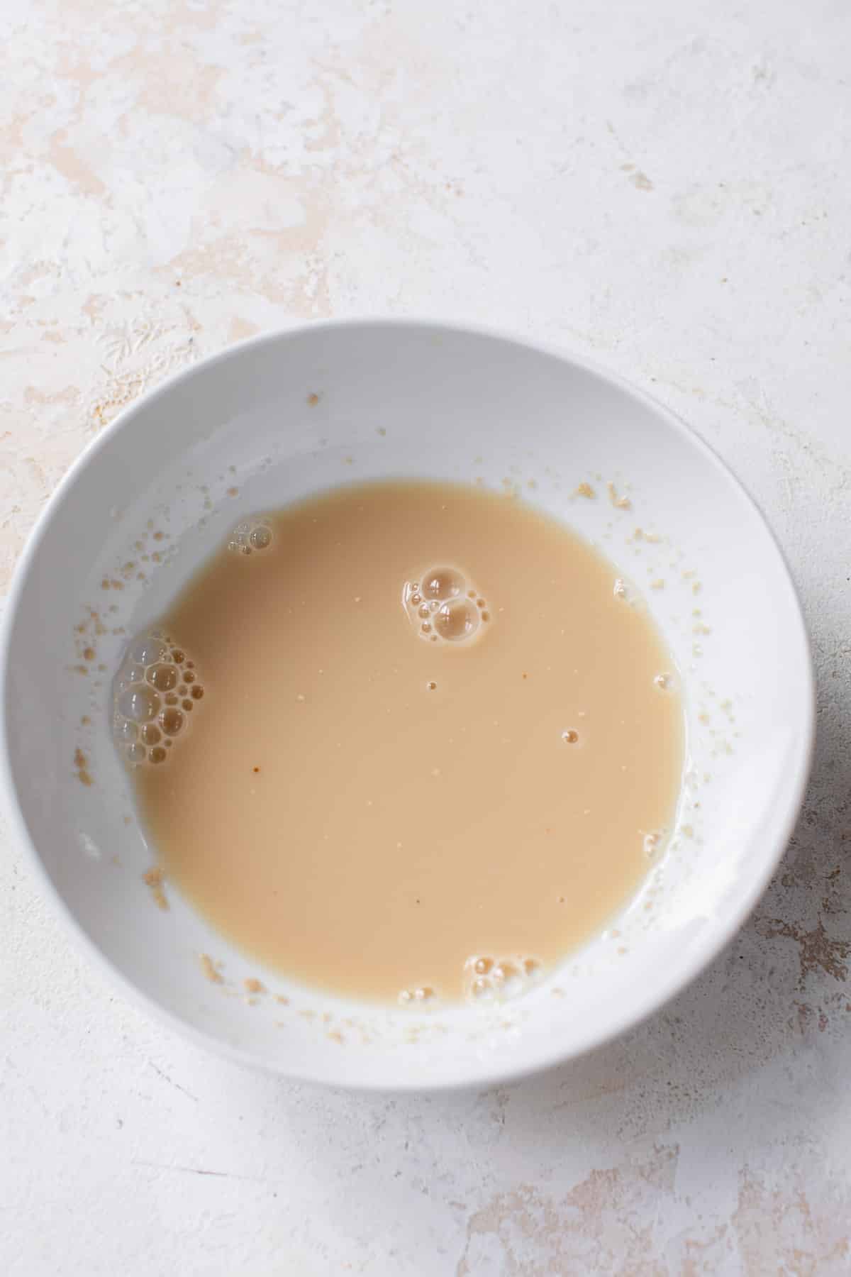 overhead view of a liquid yeast mixture for vegan cinnamon rolls in a white bowl.