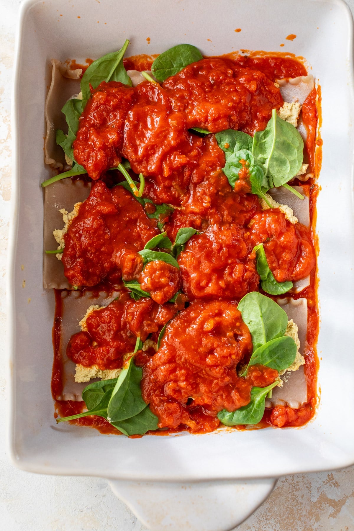 overhead view of assembling a vegan lasagna with marinara and spinach in a white baking dish.
