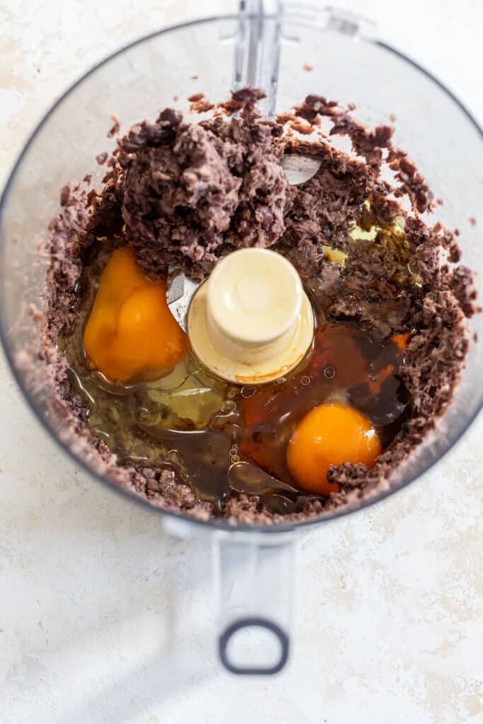 overhead view of mashed black beans and cracked eggs in a food processor.
