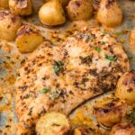 Close up on a roasted chicken breast surrounded by roasted potatoes on a sheet pan.