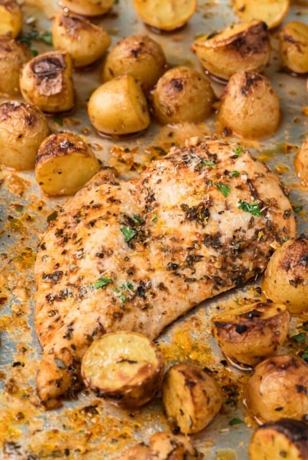 Close up on a roasted chicken breast surrounded by roasted potatoes on a sheet pan.
