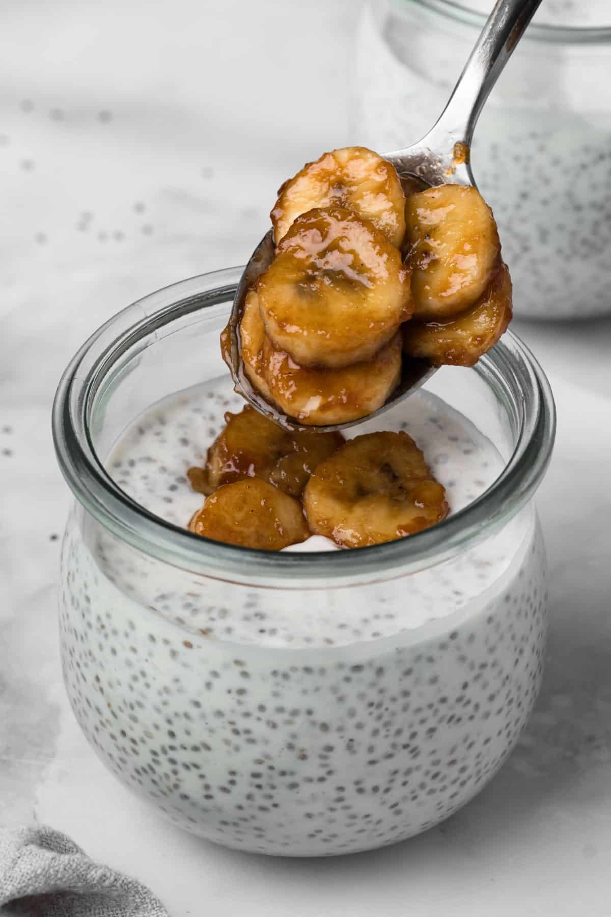 Side view of a spoon dropping caramelized bananas on top of a jar of coconut chia pudding.