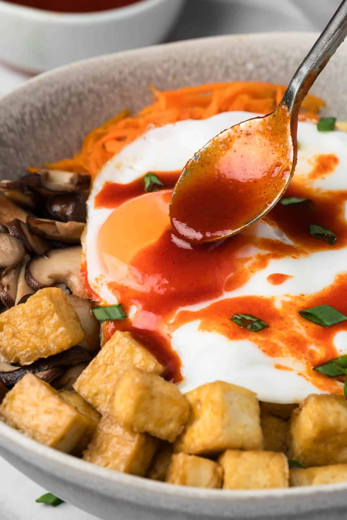 side view of a spoon drizzling red sauce over a tofu bibimbap bowl.