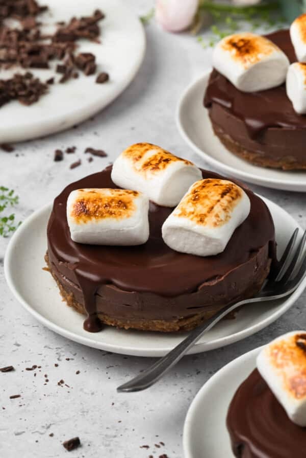 side view of 3 vegan s'mores cheesecakes on white plates with vegan marshmallows on top.