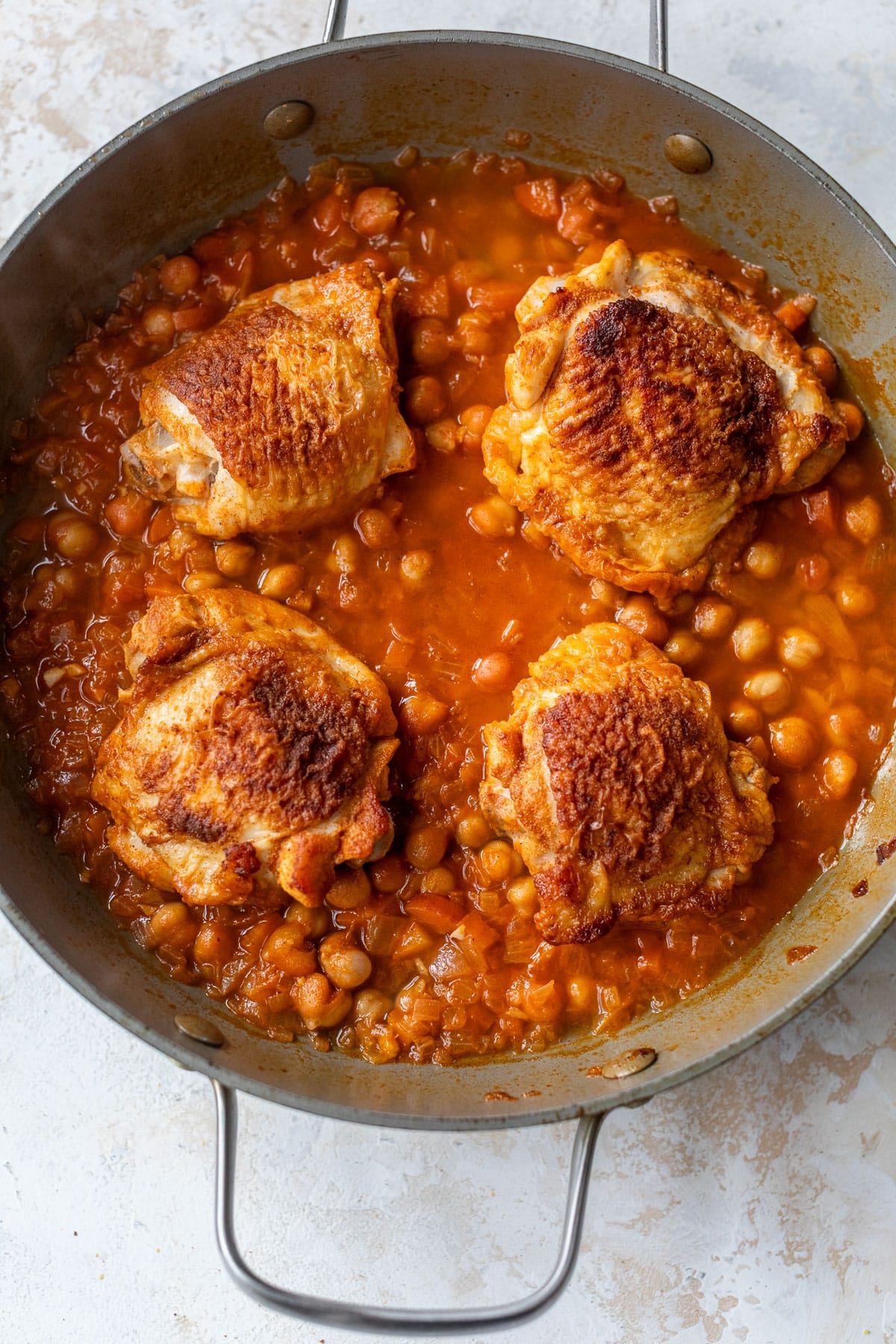 overhead view of seared chicken thighs braising in a red chickpea sauce in a skillet.
