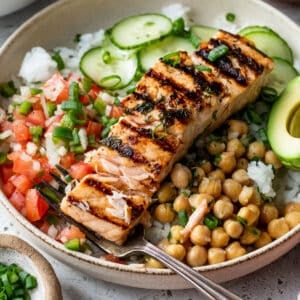 large bowl filled with rice, chickpeas, tomatoes, cucumber, avocado, and grilled salmon with a fork in the salmon