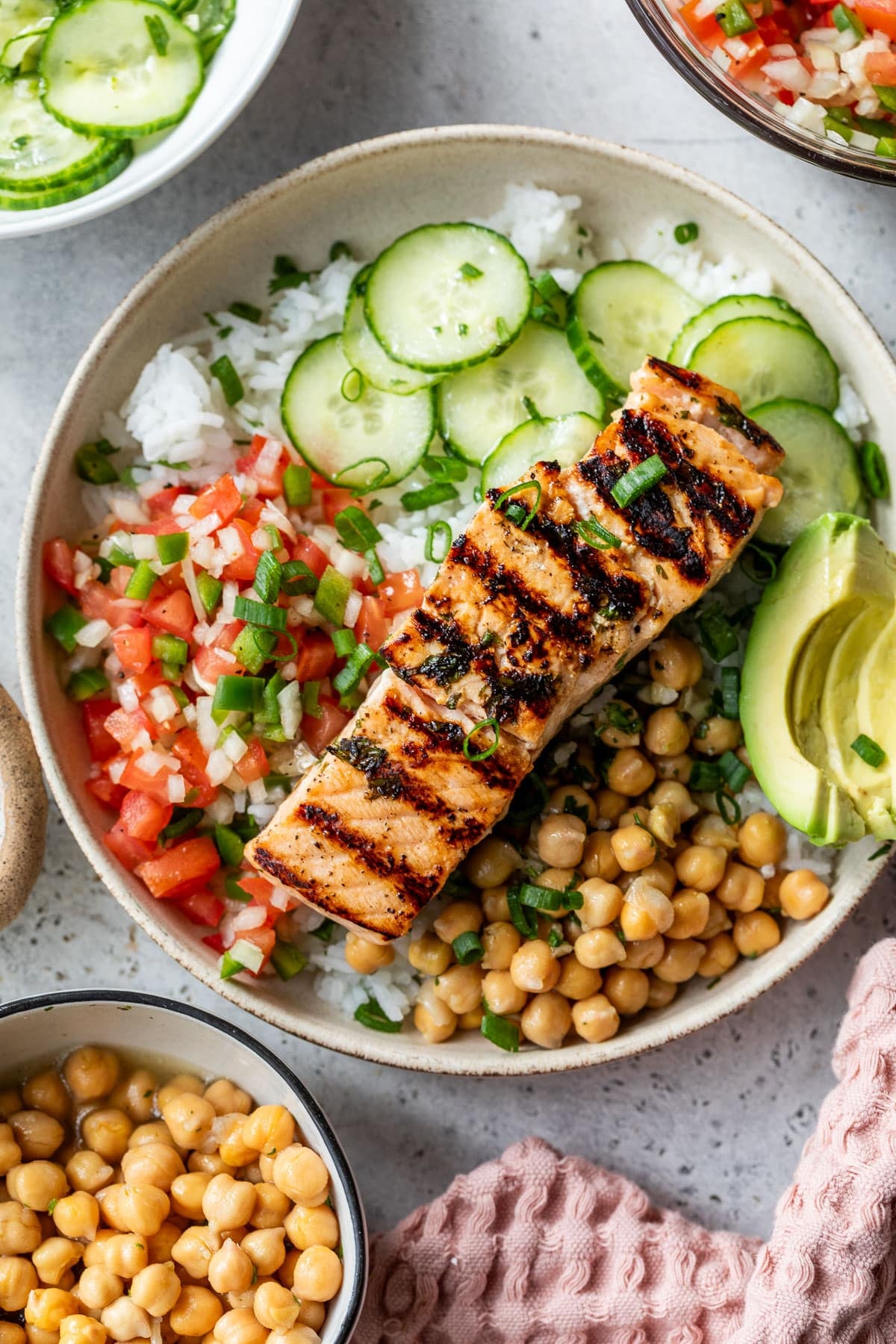 overhead shot of a bowl or rice with vegetables like cucumber and tomatoes, along with a piece of grilled salmon