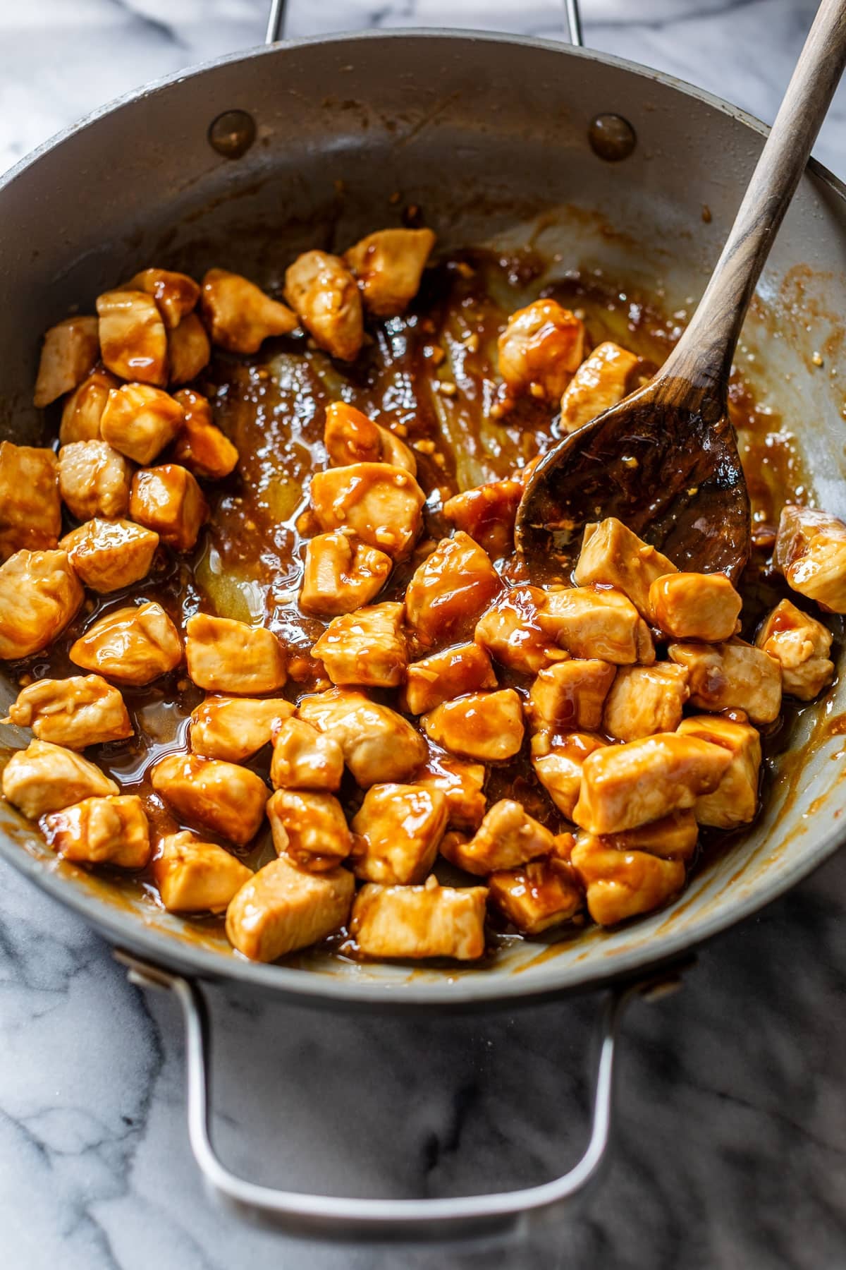 side view of a wooden spoon tossing seared chicken bites in sesame sauce in a skillet.
