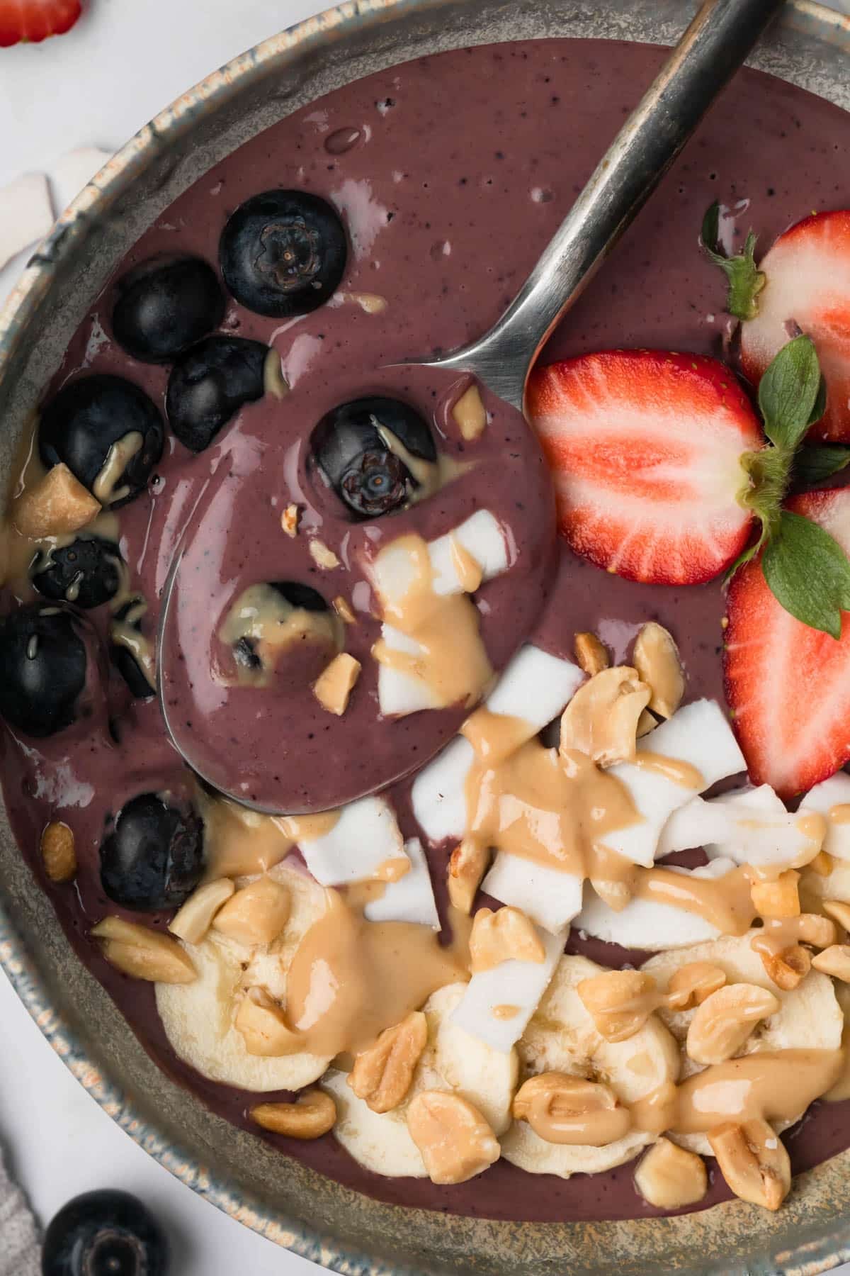 overhead view of a spoon resting in a purple peanut butter acai bowl with fruit, peanut butter, and coconut on top.