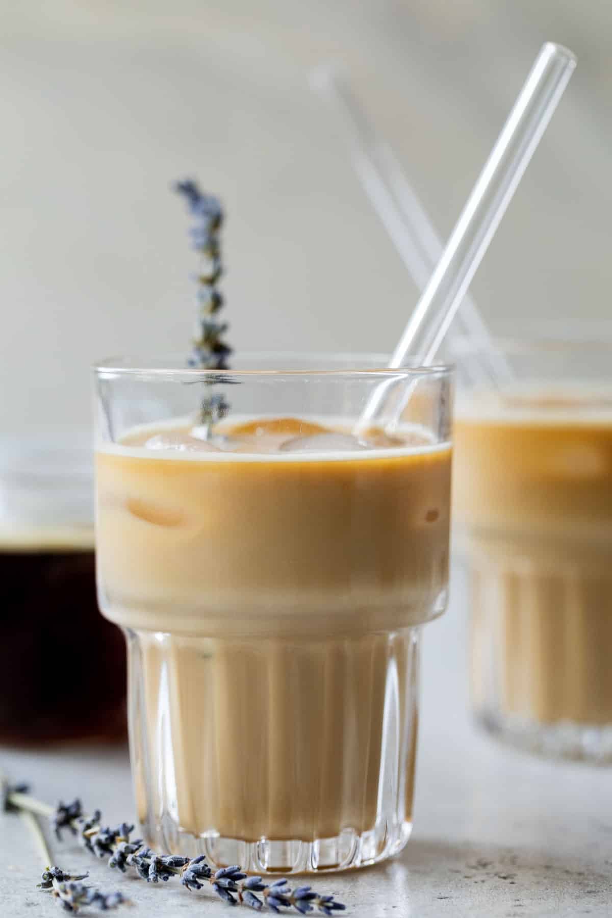 side view of two iced lavender dirty chai lattes in glasses with a glass straw and garnished with fresh lavender.