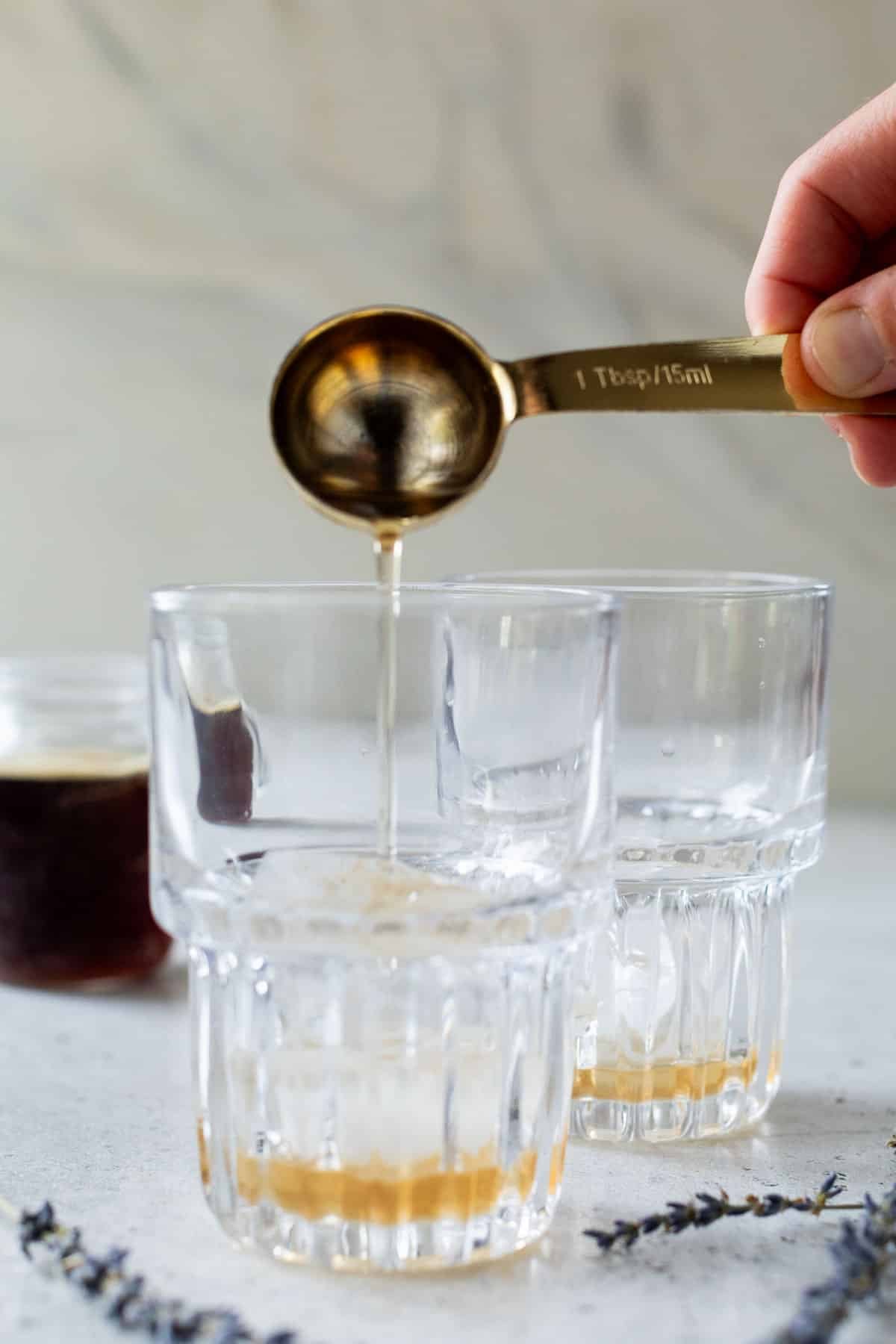 side view of a hand pouring 1 tablespoon of lavender chai syrup into a two glasses.