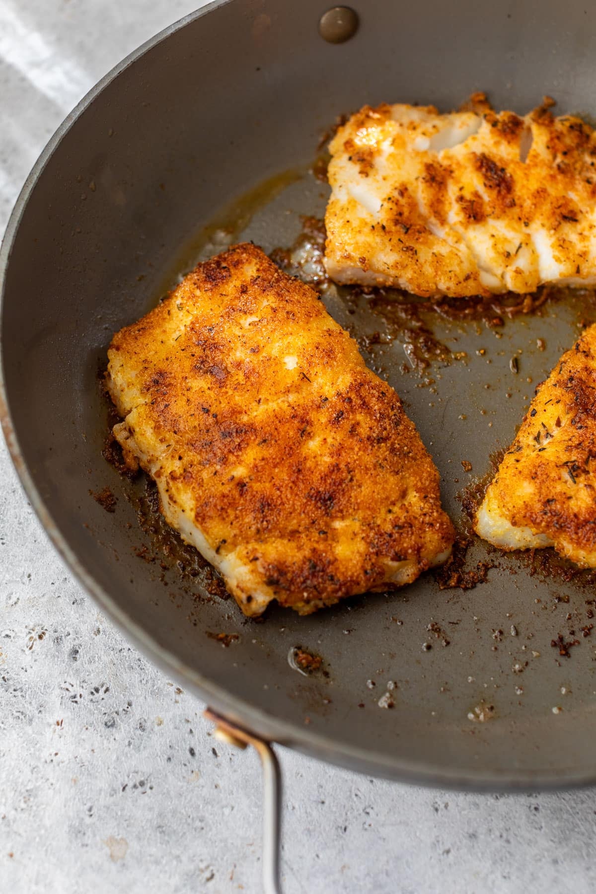 side view of 3 pieces of seasoned cod frying in a large skillet.