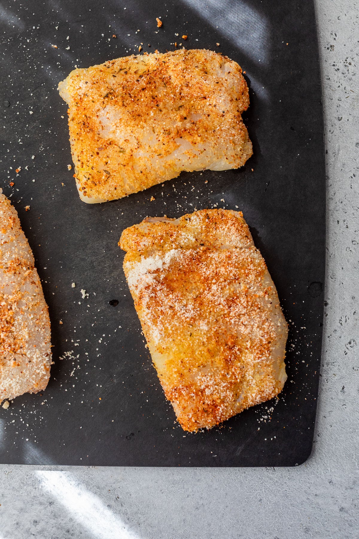 overhead view of 3 pieces of raw seasoned cod on a black cutting board.