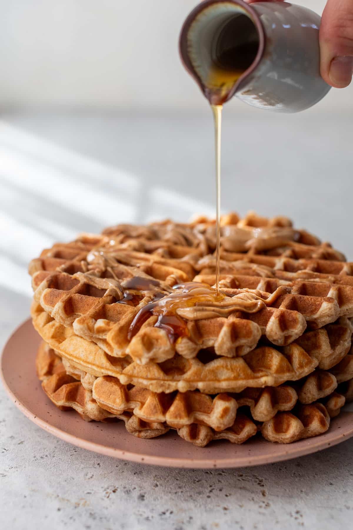 side view of a hand pouring maple syrup onto a stack of peanut butter waffles.