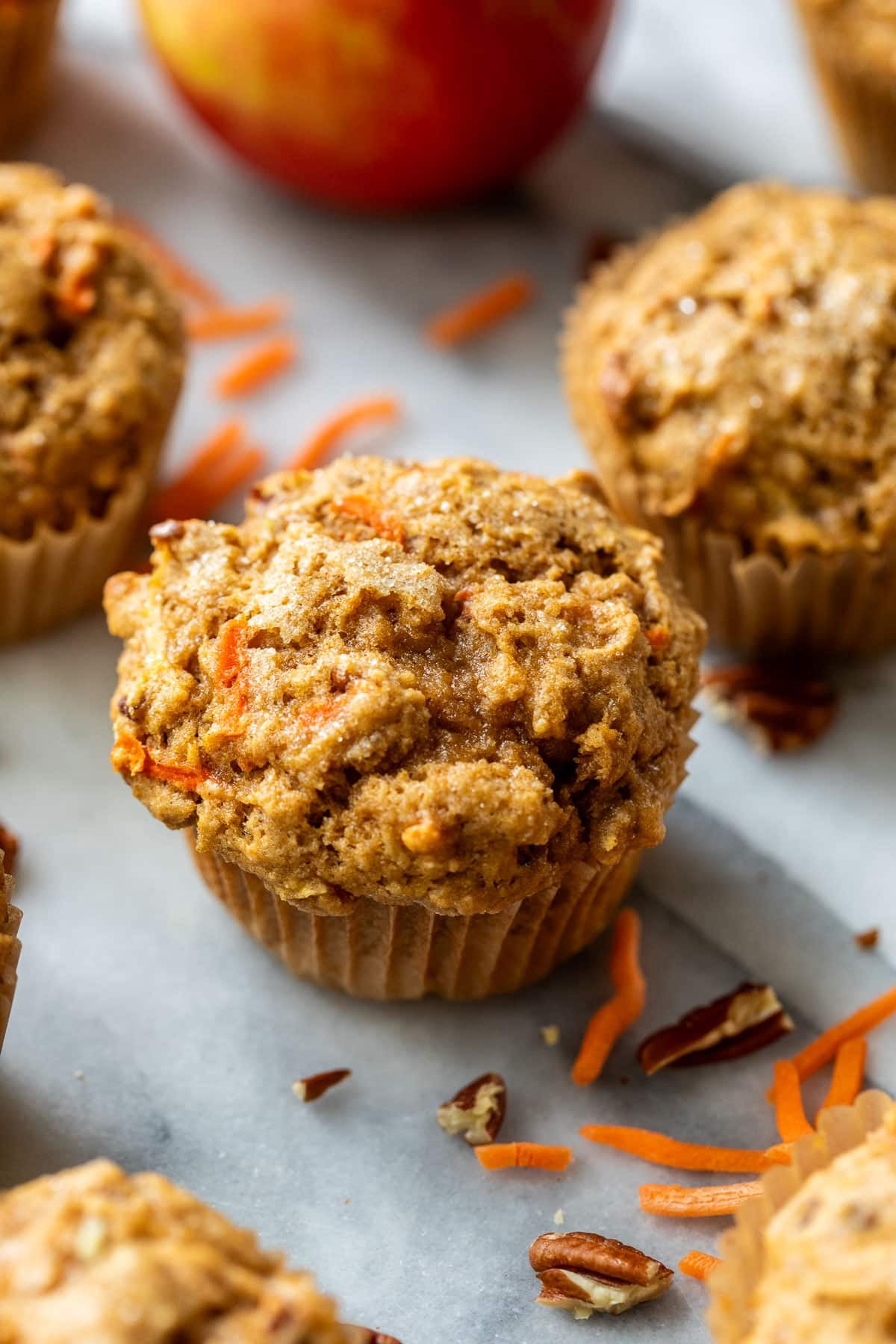 side view of baked apple carrot muffins surrounded by chopped pecans and shredded carrots.