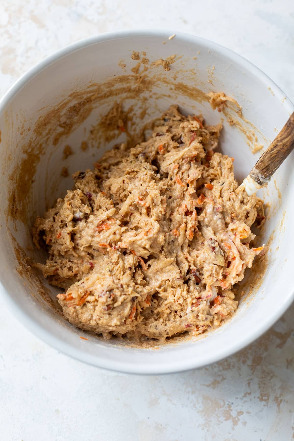 overhead view of a wooden spoon stirring the batter for apple carrot muffins in a white bowl.