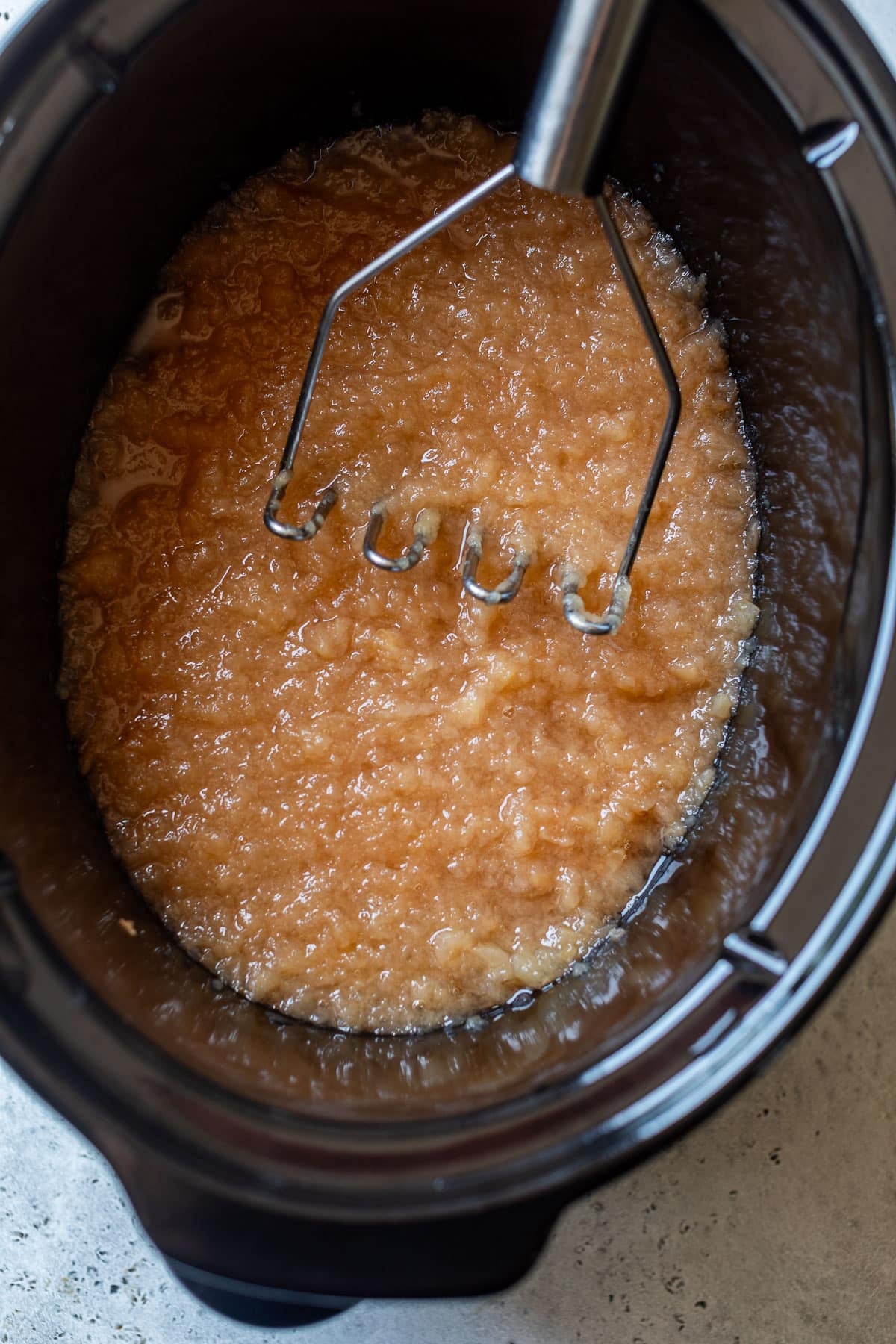 overhead view of a potato masher mashing cooked apple slices in a slow cooker.