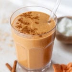 side view of a pumpkin smoothie topped with cinnamon and a glass straw in a glass.